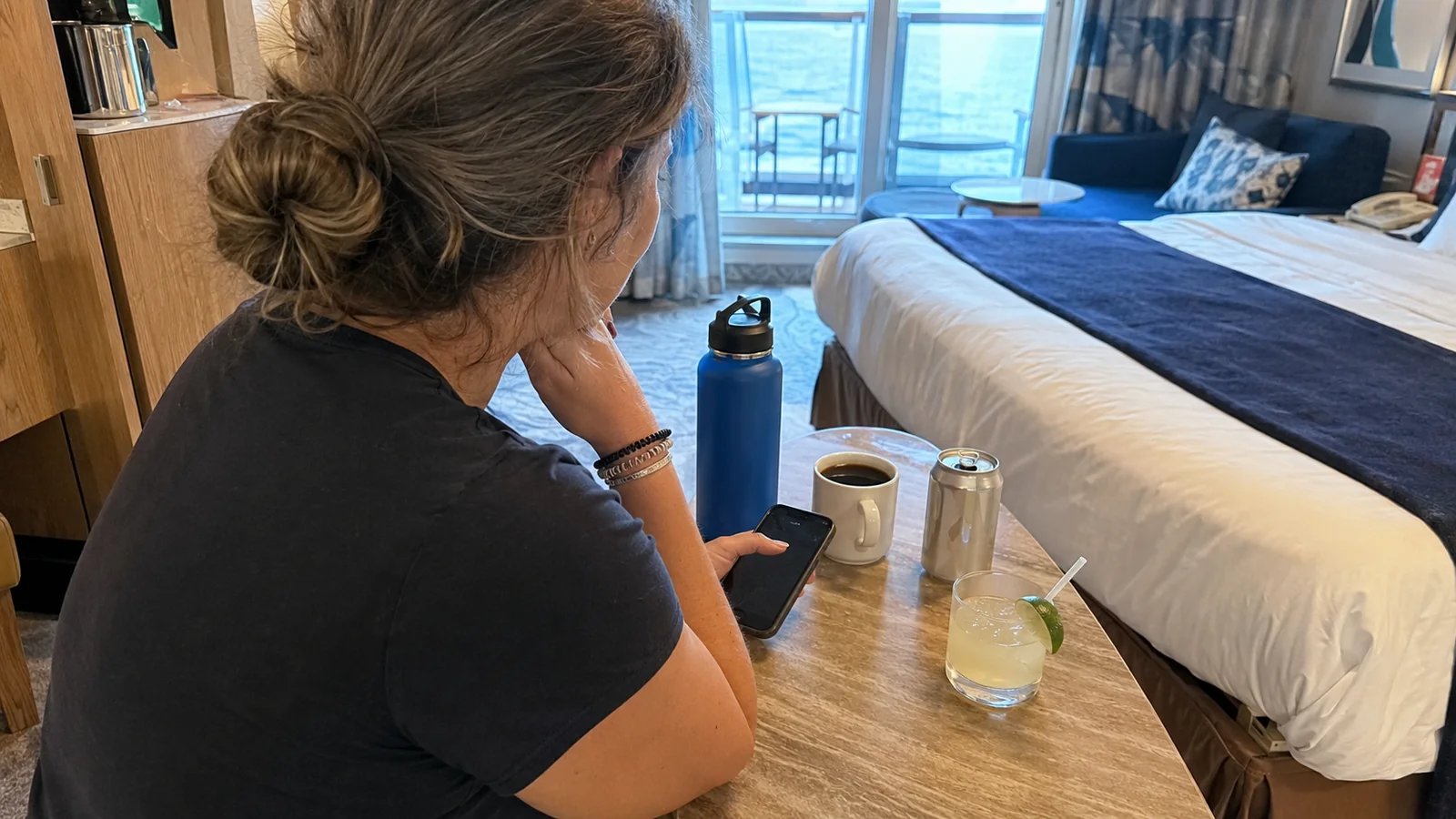 Woman seated at a cruise cabin table with a phone and drinks beside the ocean-view window