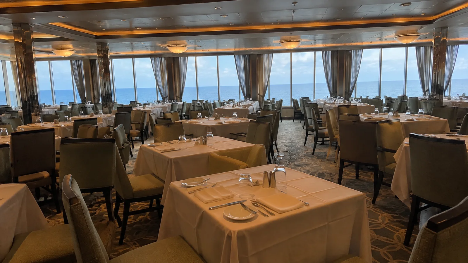 Empty dining tables are set beside panoramic ship windows.