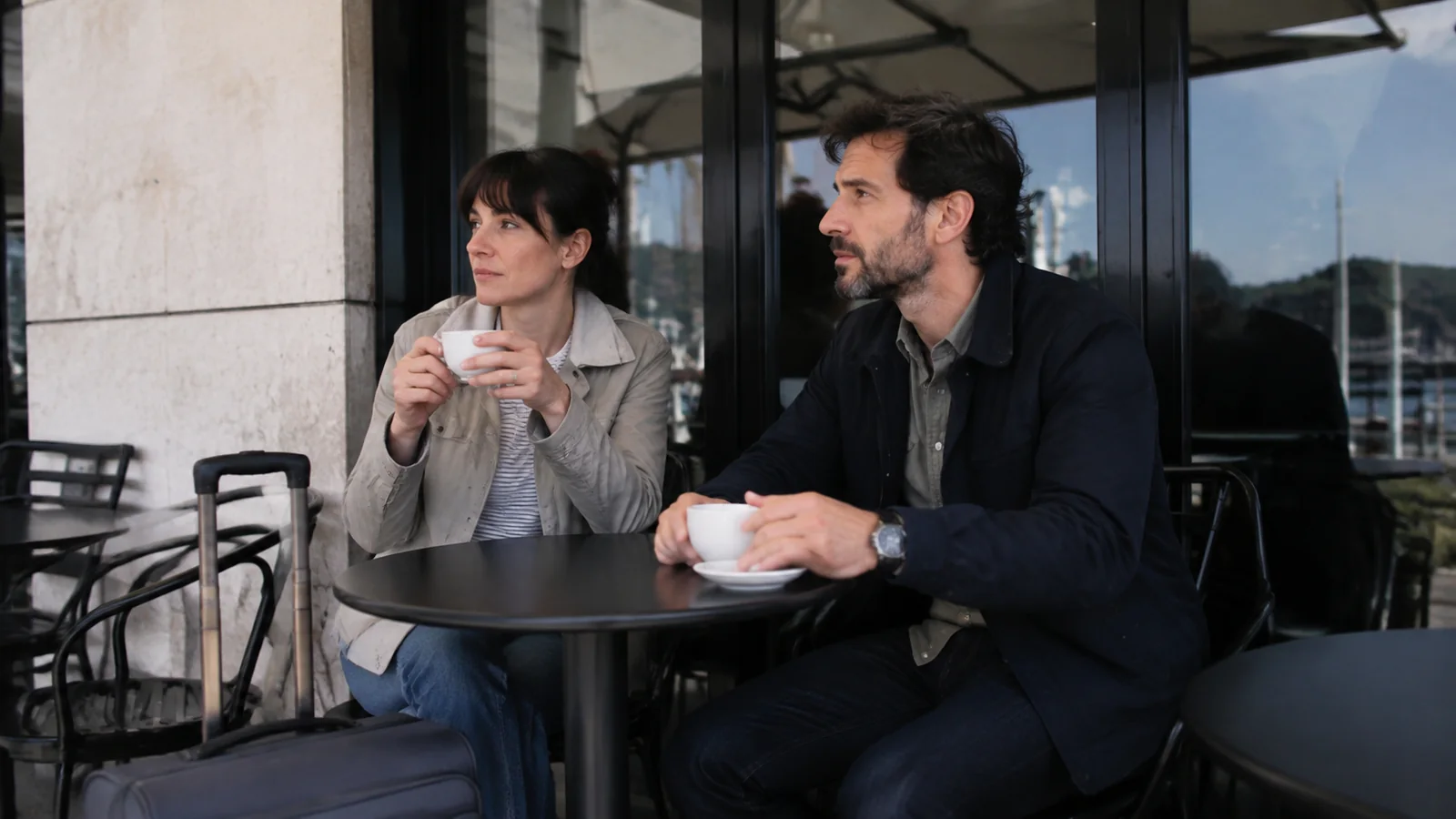 Two travelers sit at an outdoor café table with coffee cups.