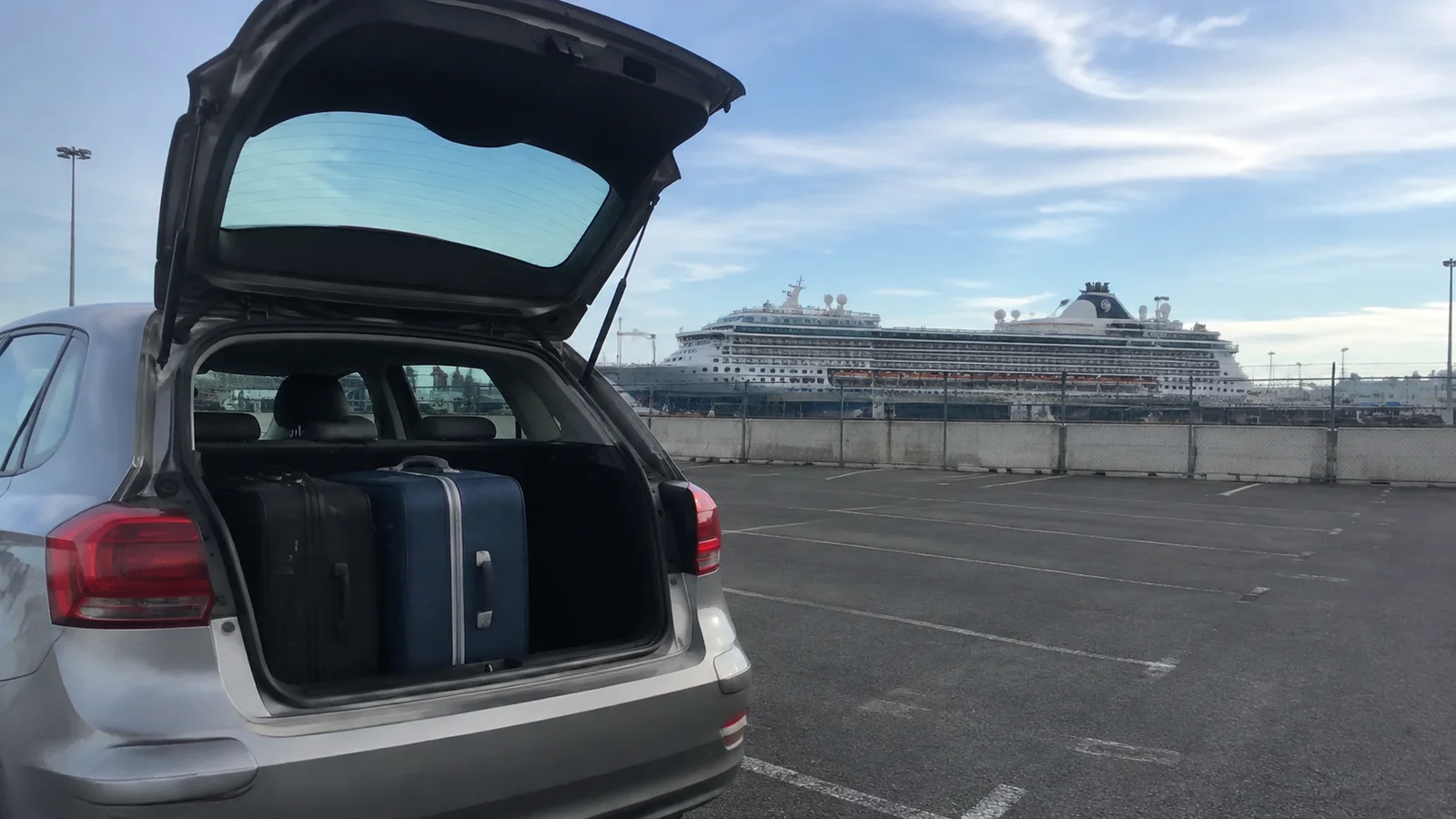 Open car trunk holds suitcases with a cruise ship across the port.