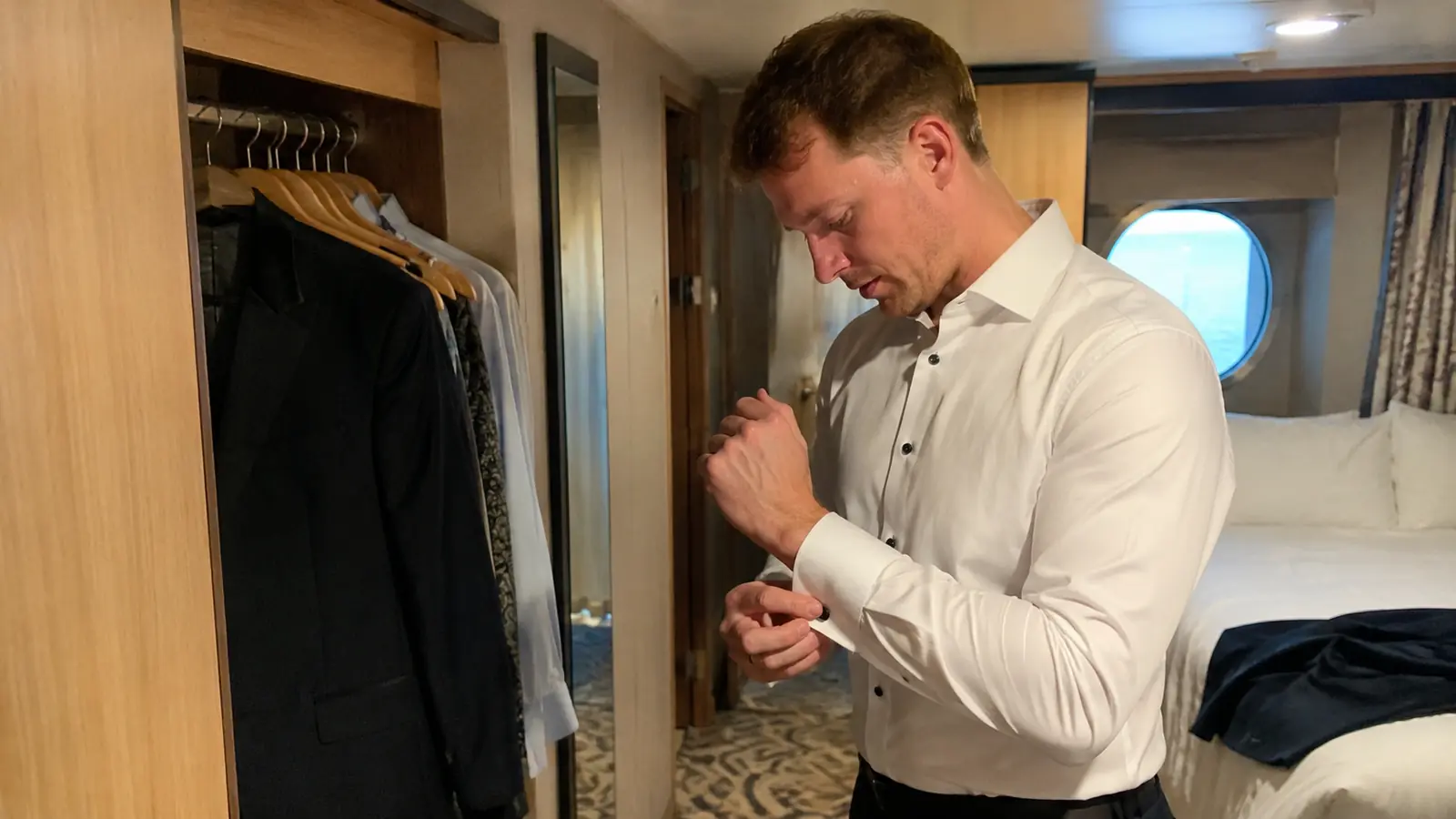 Man fastening his shirt cuff beside a dark suit hanging in a cruise cabin