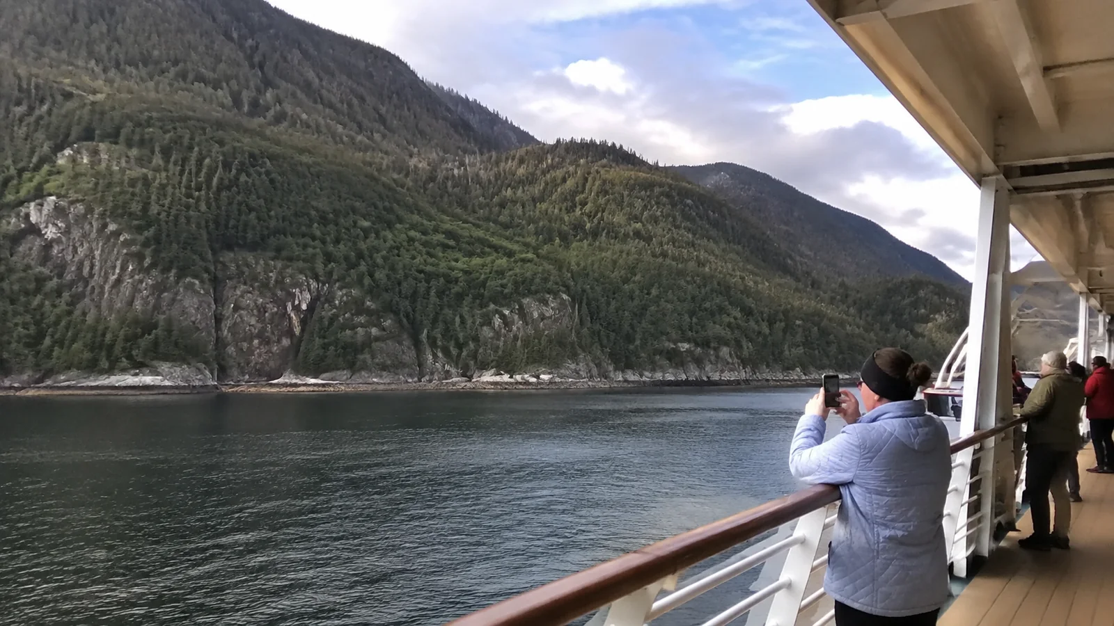Passengers photographing a forested mountain coast from a ship.