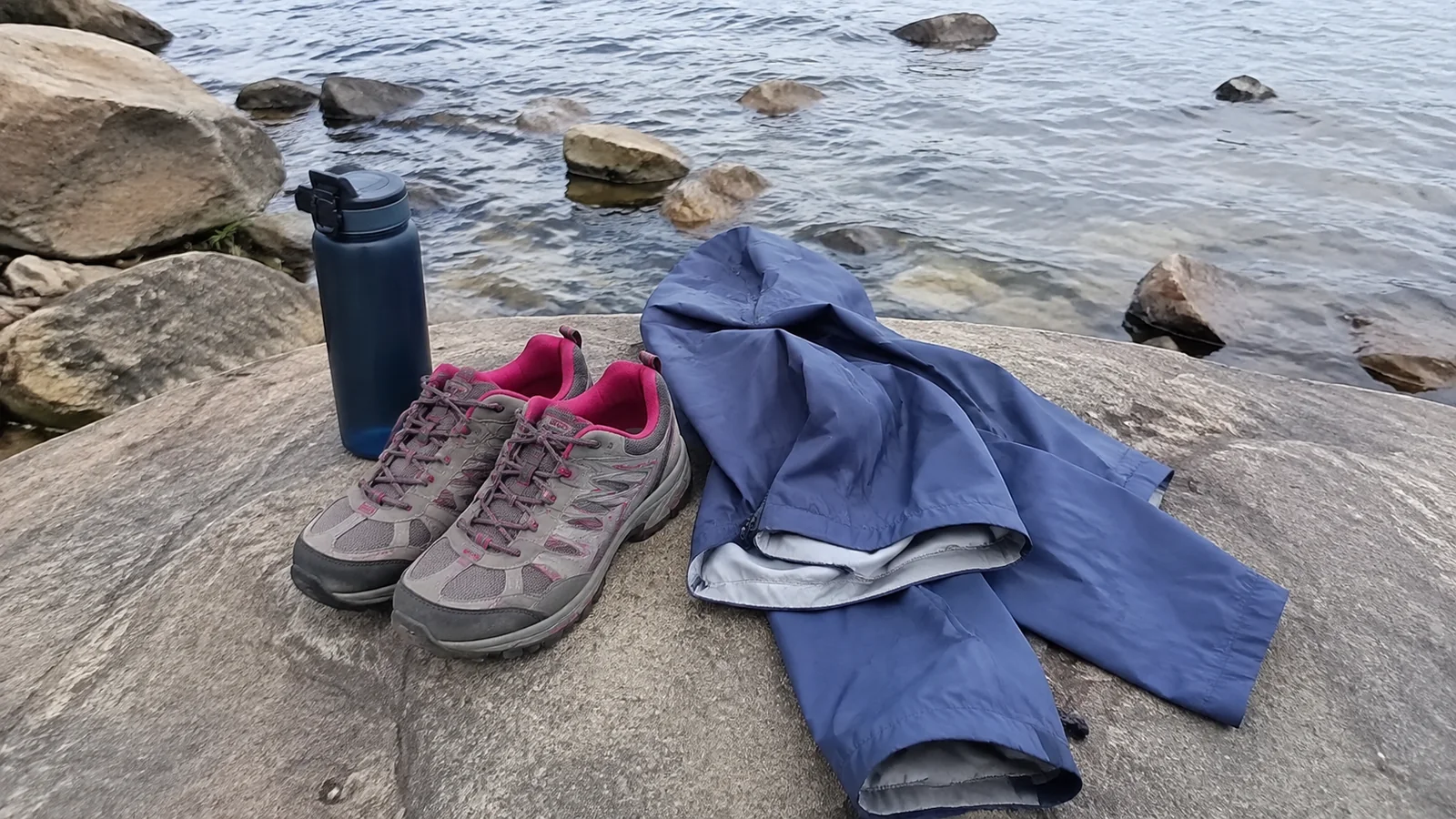 Walking shoes, jacket and reusable bottle rest on a lakeside rock.