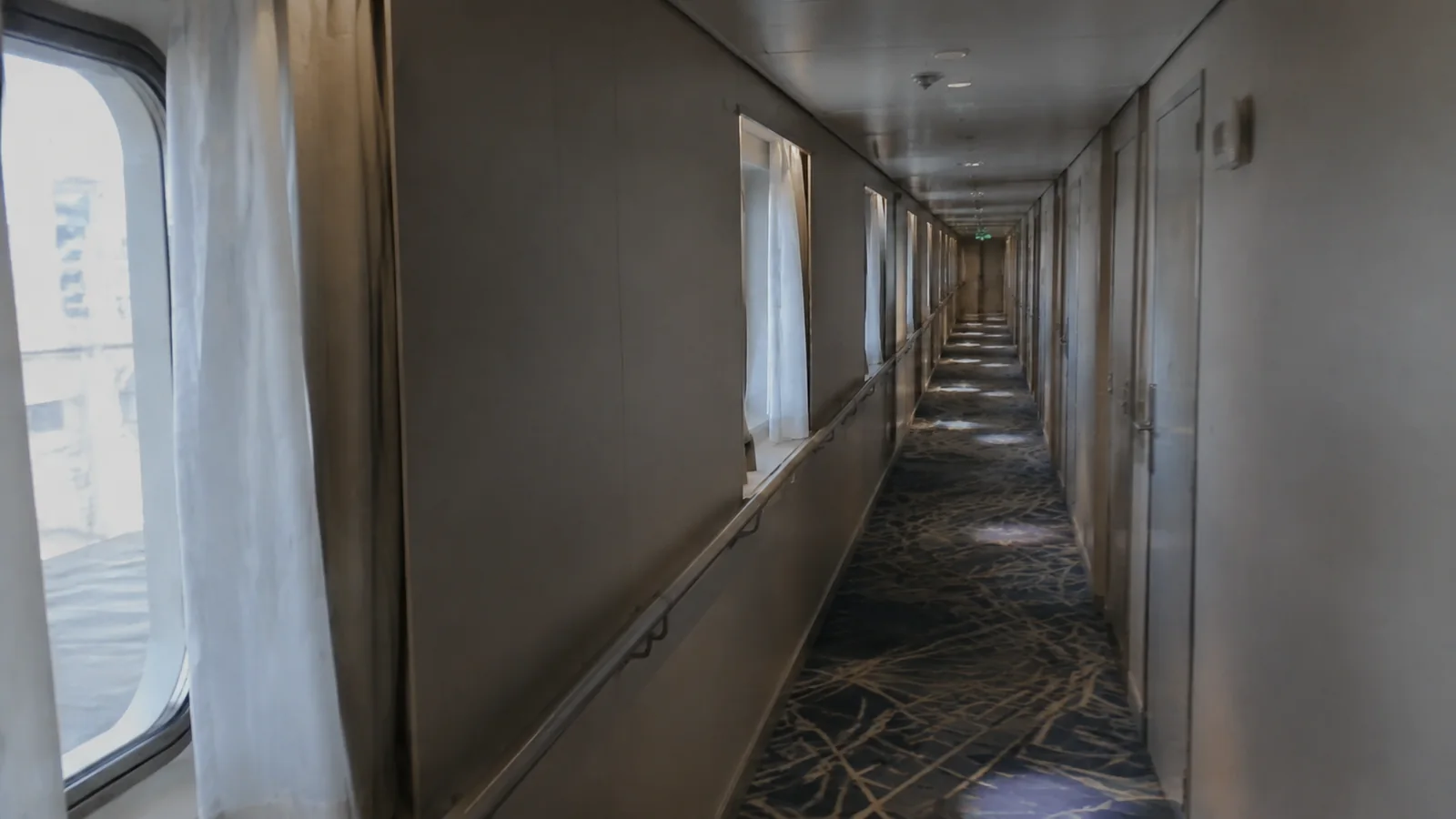 Long empty corridor runs beside windows and closed stateroom doors.