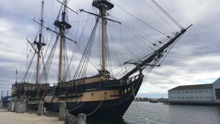 Historic sailing ship moored beside a quay with tall masts and rigging.