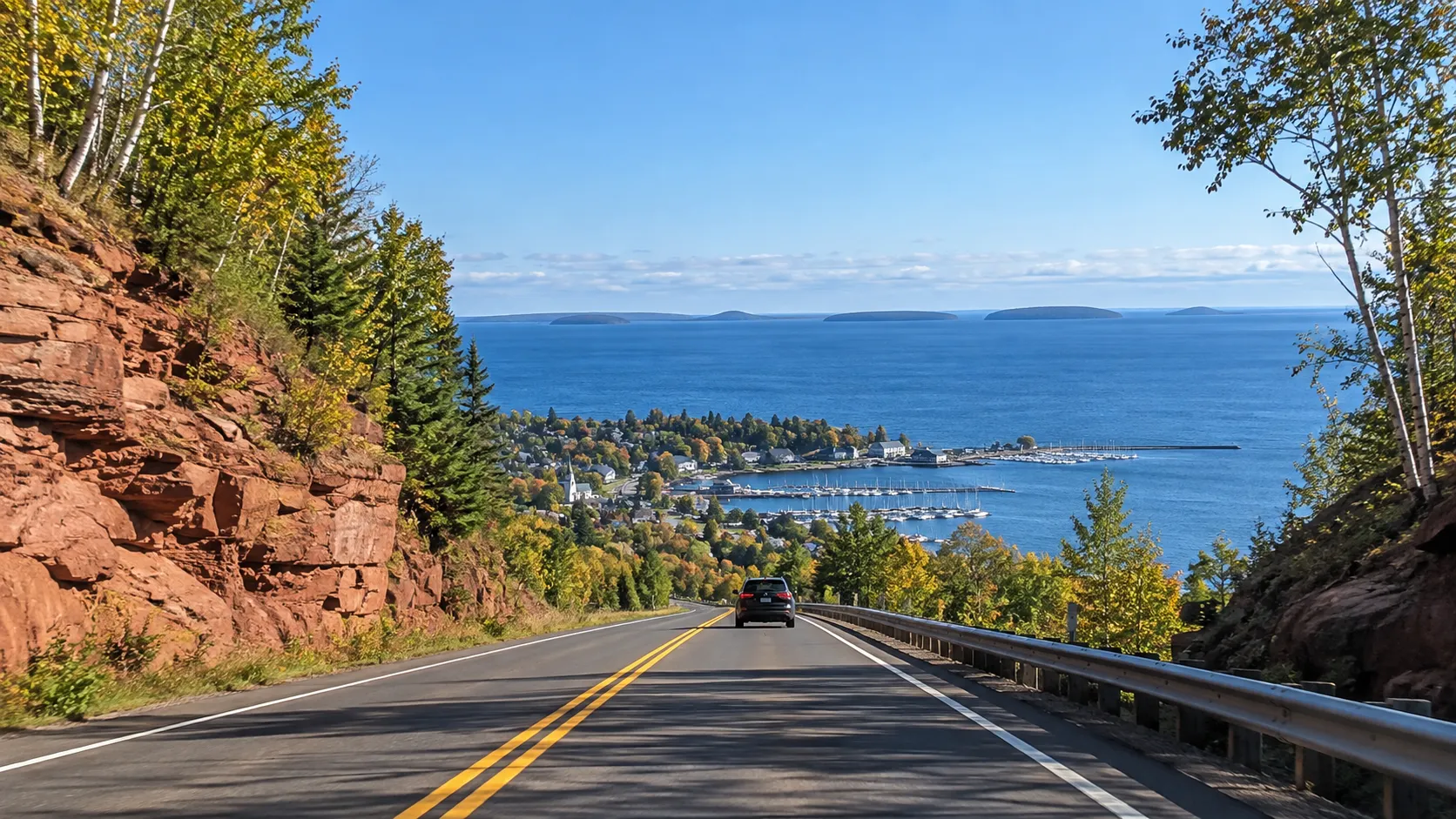 A lakeshore road descends between red sandstone and birch trees toward Bayfield and the Apostle Islands.