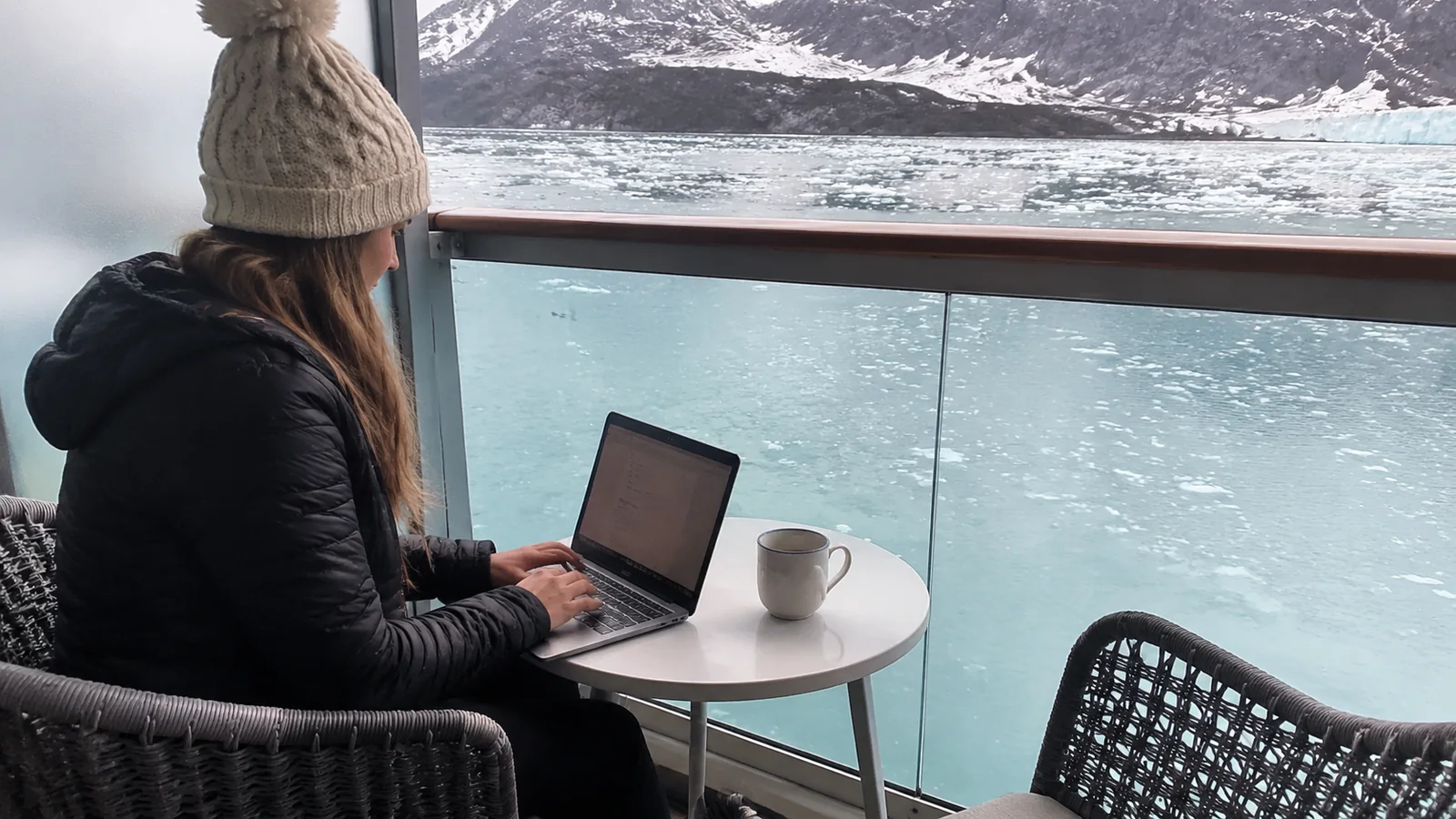 Woman using a laptop on a cruise ship balcony with a glacier behind her
