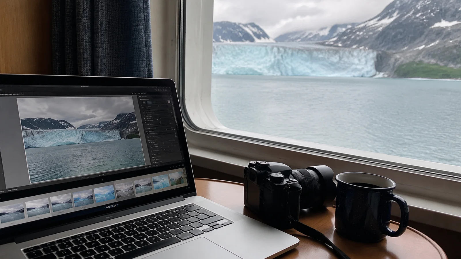 Laptop displaying a glacier photo beside a camera and mug with a glacier outside the window