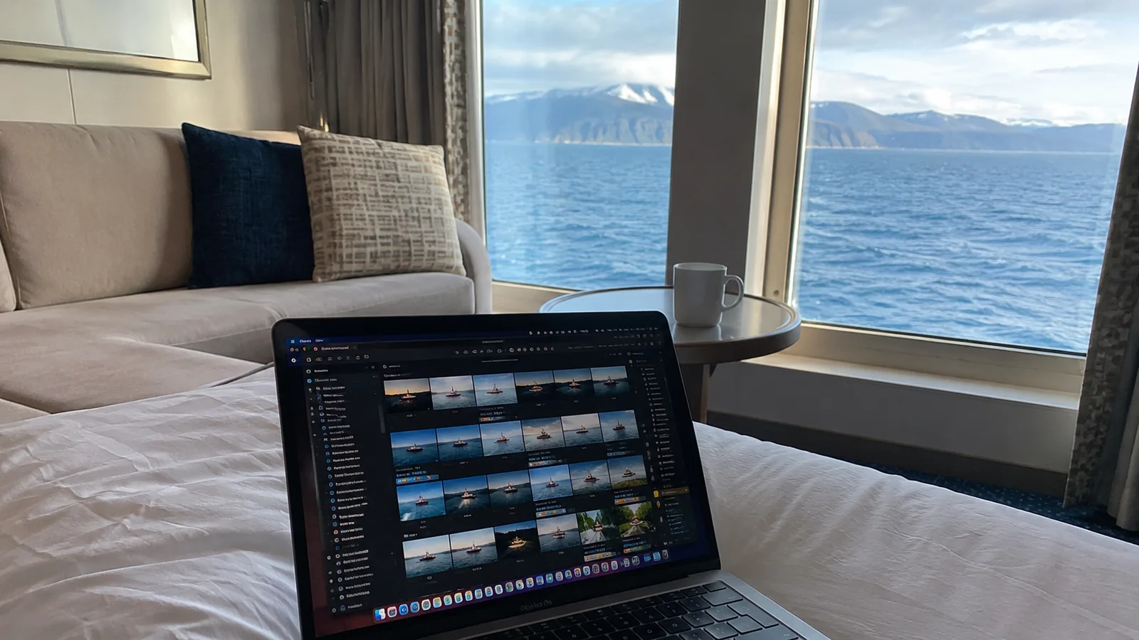 Laptop displaying a photo library on a cruise cabin bed beside an ocean-view window