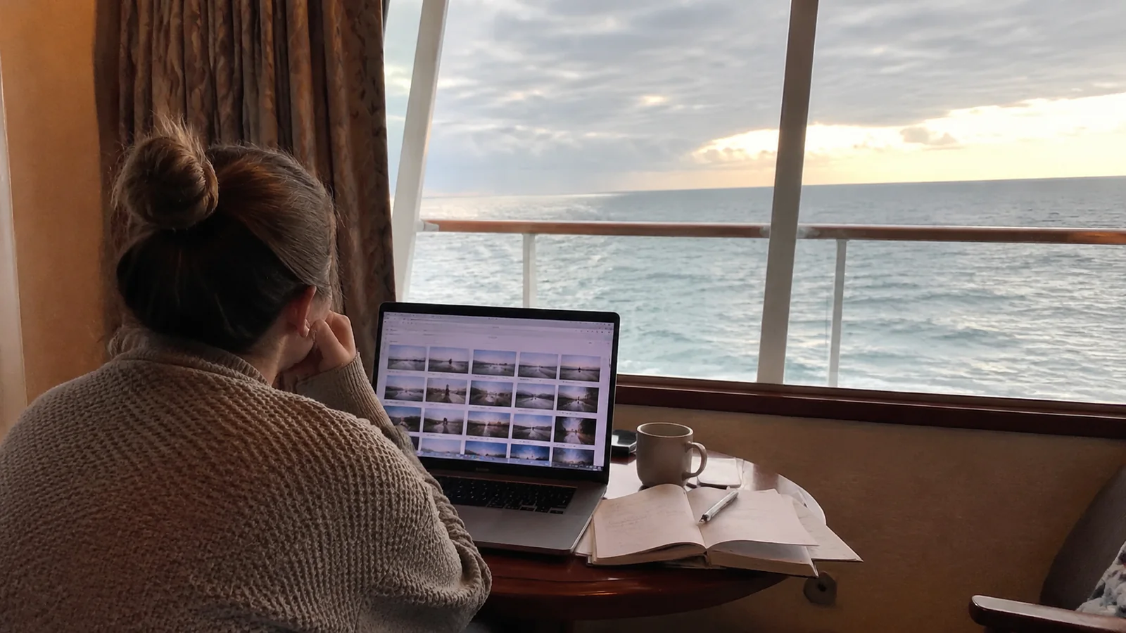 Woman reviewing image thumbnails on a laptop at a cabin desk overlooking the sea