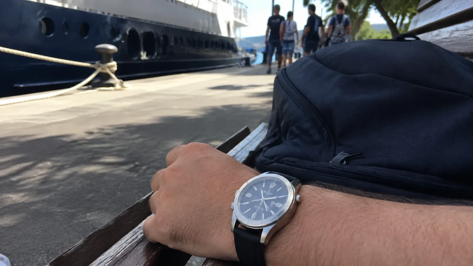 Traveler checking a wristwatch beside a backpack at a pier.