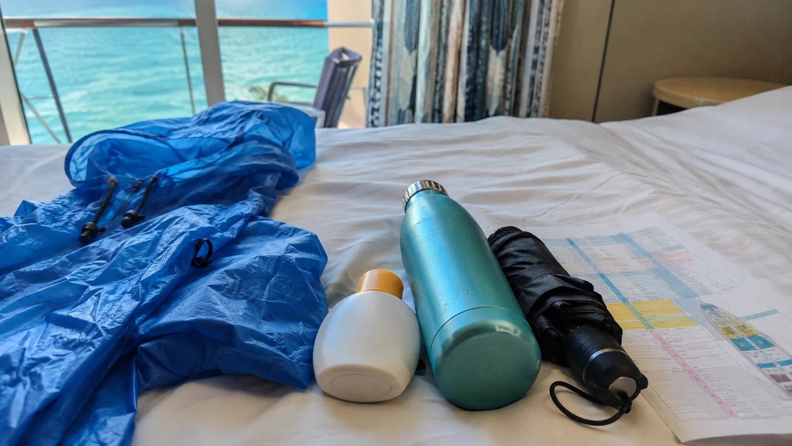 Rain jacket, sunscreen, water bottle, umbrella, and map arranged on a cruise cabin bed